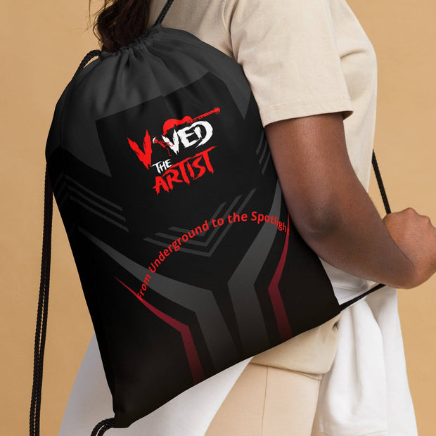 Close-up of Viv'ed the Artist drawstring bag featuring bold red and gray design with text, worn as a backpack by a woman in beige outfit.